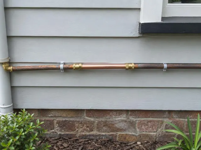 Repaired copper water pipe on an exterior weatherboard wall above a brick base in Waverley